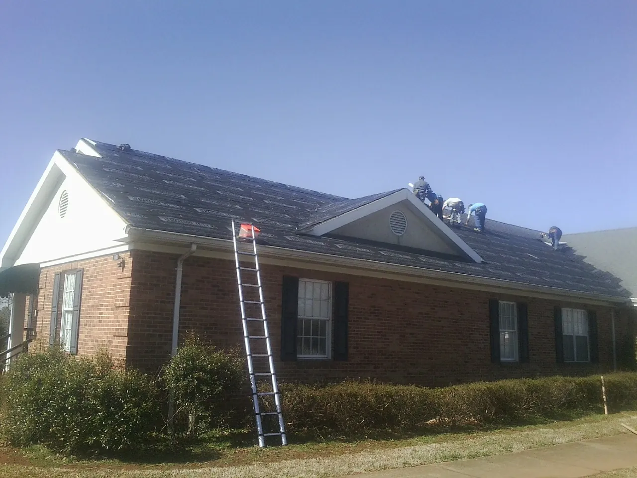 Crew tearing shingles off a brick ranch roof with a ladder set — Emergency Roof Repair work by our crew in Banco