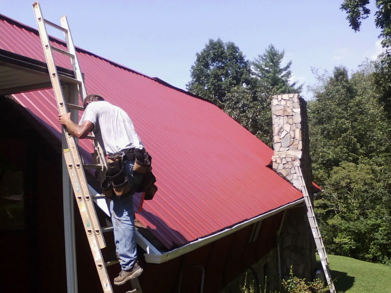 Roofer on a ladder beside a red metal roof and stone chimney — Soffit & Fascia Repair by Roofing Brothers in Banco